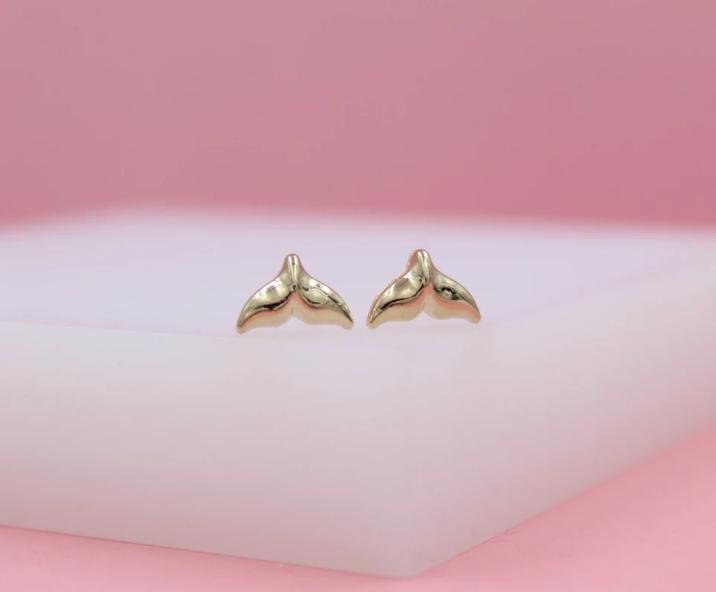 A pair of shiny gold whale tail stud earrings rests on a white display block against a soft pink background.