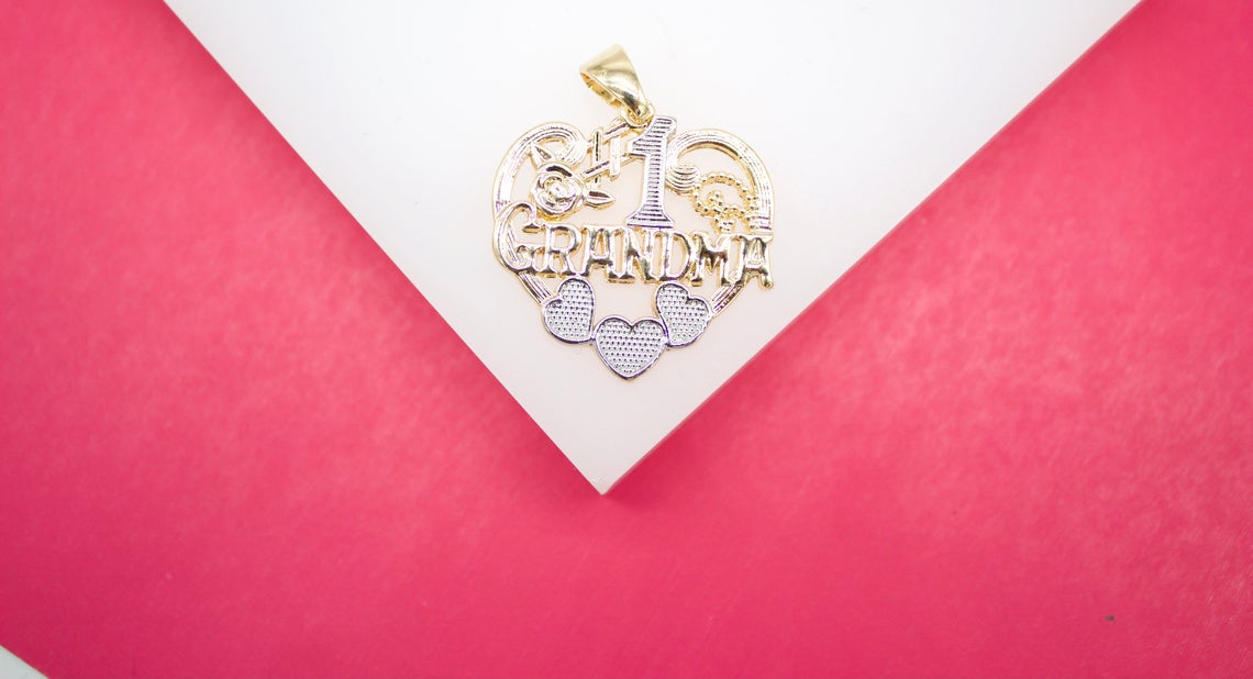 A gold "#1 Grandma" pendant with heart accents rests on a white display against a vibrant pink background.