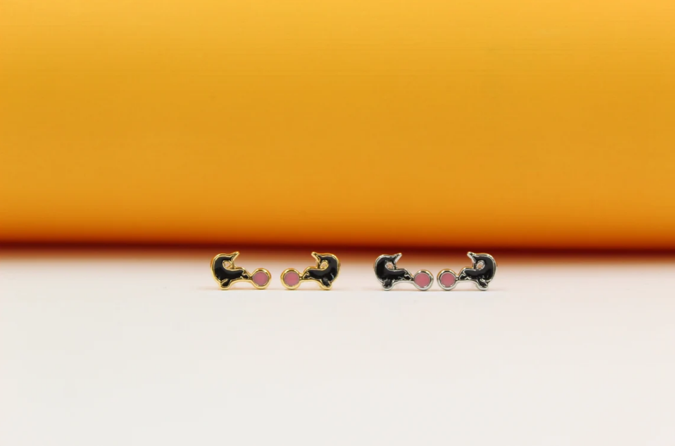 Two pairs of gold and silver cat stud earrings feature playful black enamel bodies and cute pink accent circles against a bright orange and white backdrop.