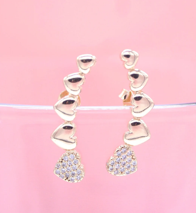 A pair of gold heart-shaped crawler earrings sparkle against a soft pink background.