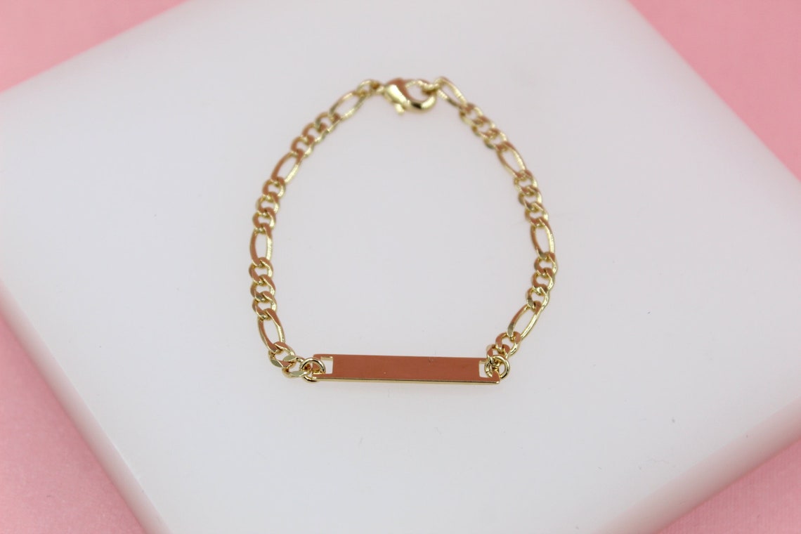 A delicate gold ID bracelet rests on a white display block against a soft pink background.