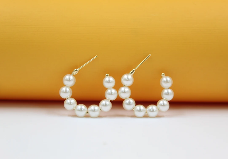 A pair of gold hoop earrings adorned with alternating white pearls sits against a warm orange and white backdrop.