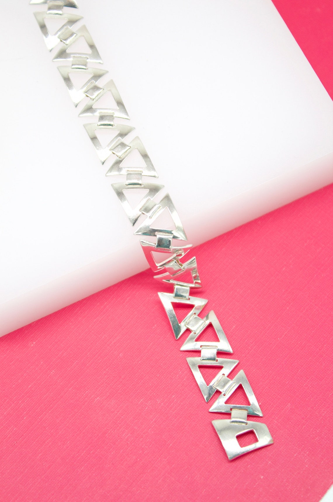 A sterling silver link bracelet featuring geometric open triangle designs rests diagonally across a white and pink background.