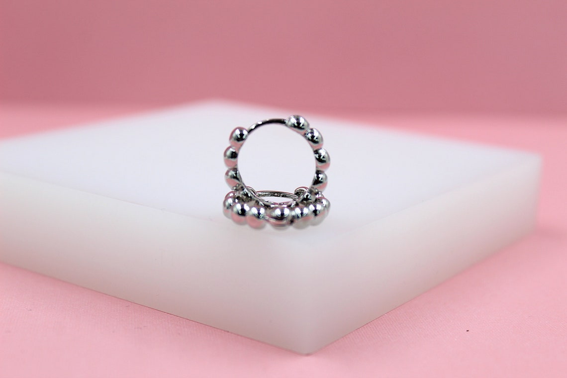A silver beaded hoop earring rests on a white display block against a soft pink background.