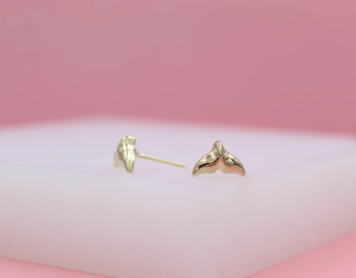 A pair of shiny gold whale tail stud earrings rests on a white display surface against a soft pink background.