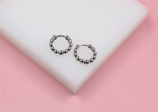 A pair of silver beaded huggie hoop earrings rests on a white block against a soft pink background.