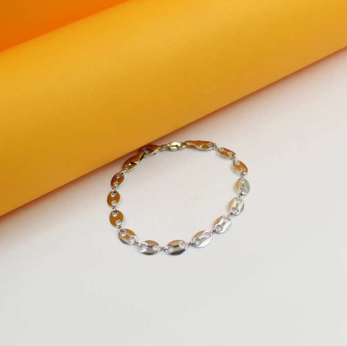 A delicate silver coffee bean link bracelet rests on a white surface against an orange backdrop.