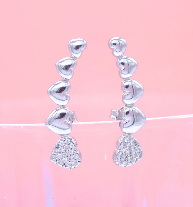 A pair of silver heart-shaped crawler earrings resting on a pink background.