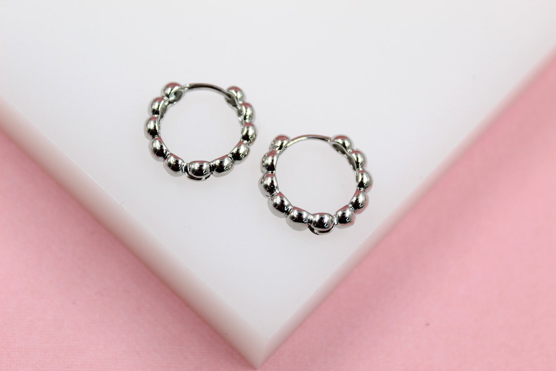 A pair of silver beaded hoop earrings rests on a white display block against a soft pink background.