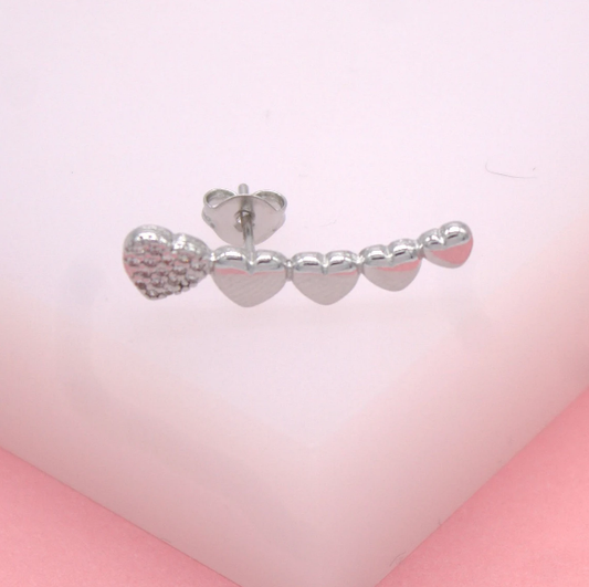 A silver ear crawler earring featuring a graduating line of sparkling stone-paved and polished metal hearts rests on a soft pink display surface.