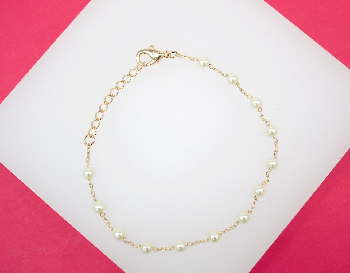 A delicate gold chain bracelet features alternating pearls and rests against a white and bright pink background.