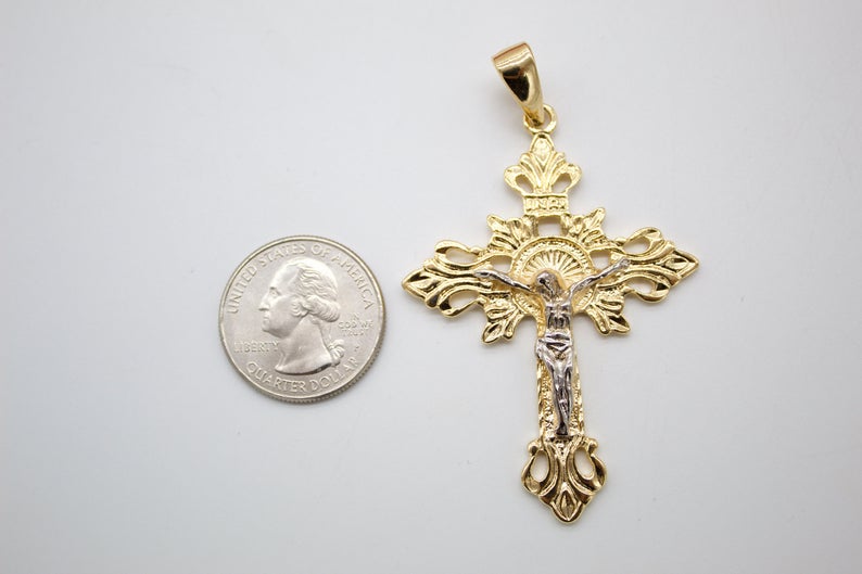 A detailed gold and silver crucifix pendant lies next to a US quarter coin for scale on a light gray surface.