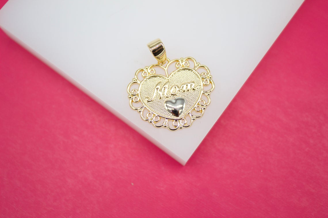 A gold heart-shaped pendant featuring the word "Mom" sits on a white surface against a vibrant pink background.