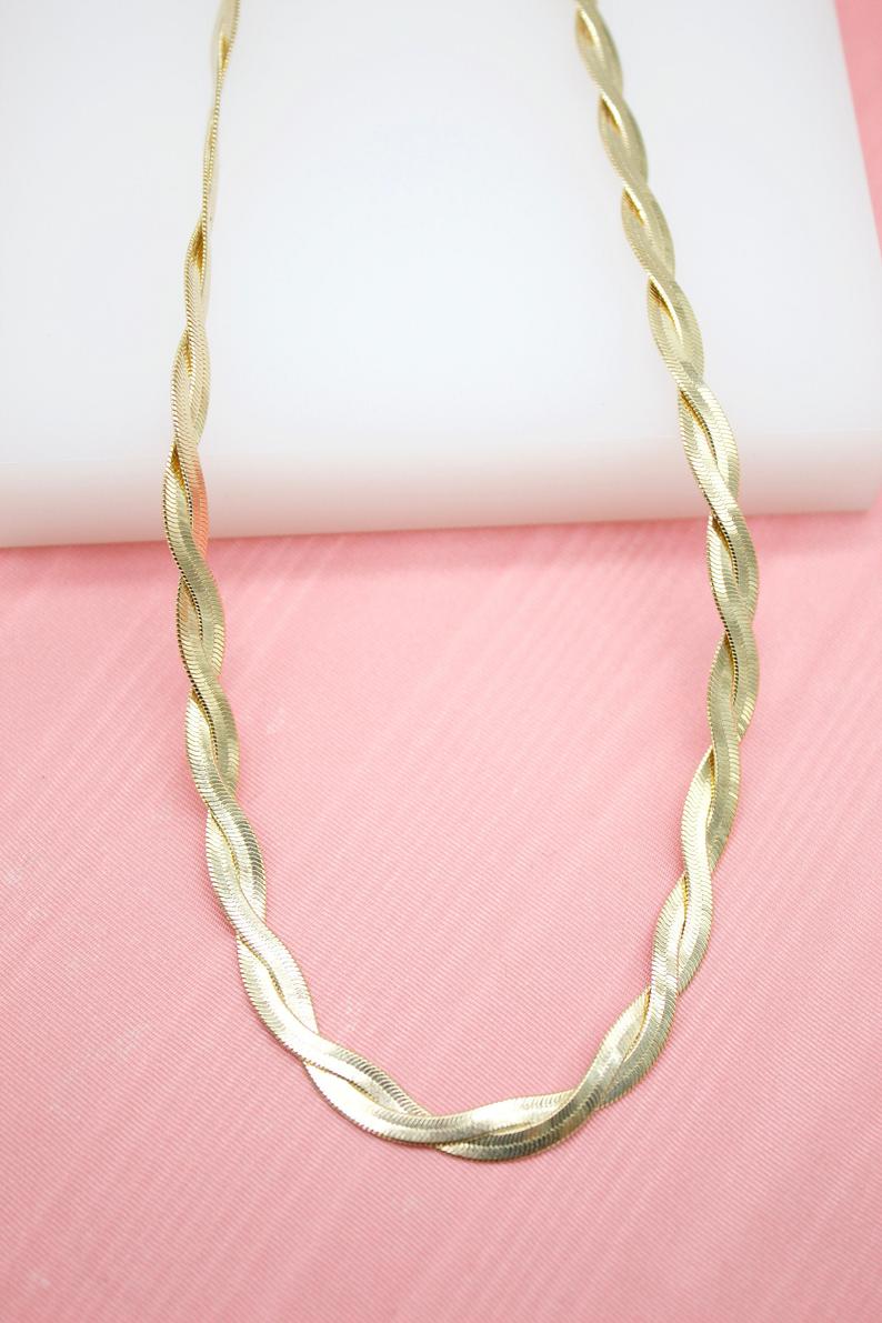 A shiny gold herringbone chain necklace drapes against a white and pink background.