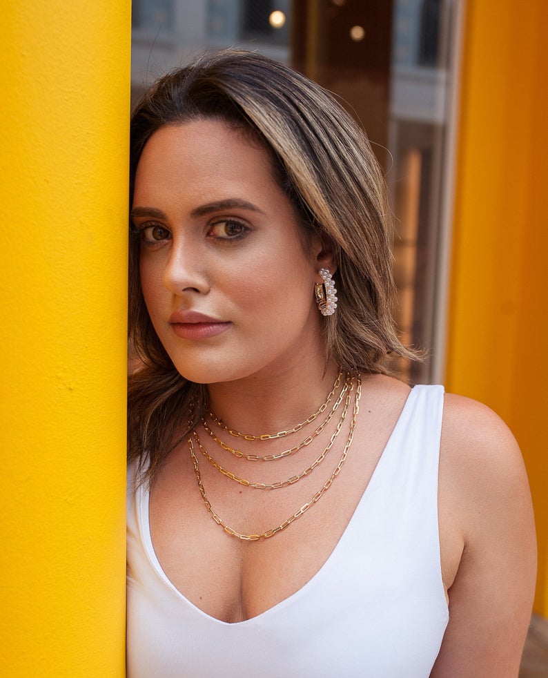 A stylish woman wearing layered gold chain necklaces and pearl hoop earrings poses next to a bright yellow pillar.