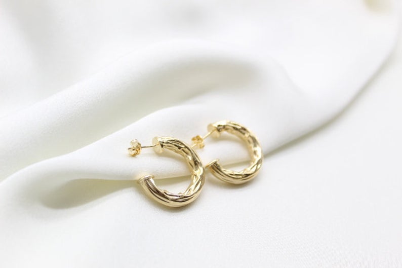 A pair of textured gold hoop earrings rests delicately on a soft white silk fabric.