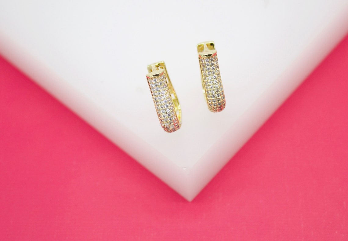 A pair of pave-set cubic zirconia gold huggie hoop earrings rests on a white display against a vibrant pink background.