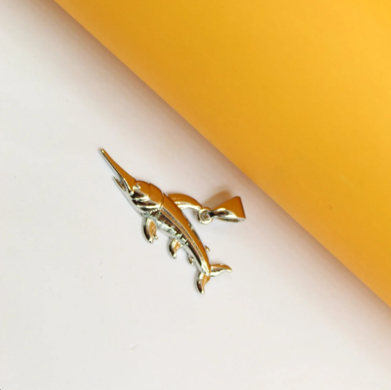 A shiny silver marlin fish pendant rests on a dual-toned white and yellow background.