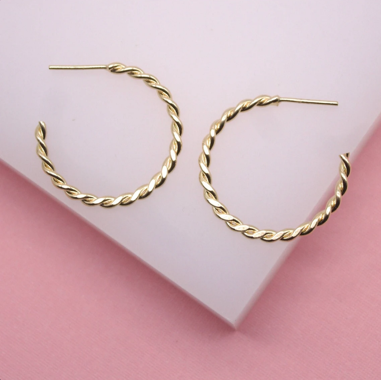A pair of elegant gold twisted hoop earrings rest on a white and pink display stand.
