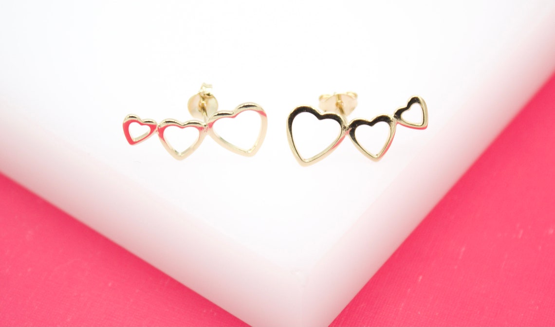 A pair of gold heart crawler earrings rests on a white display block against a vibrant pink background.