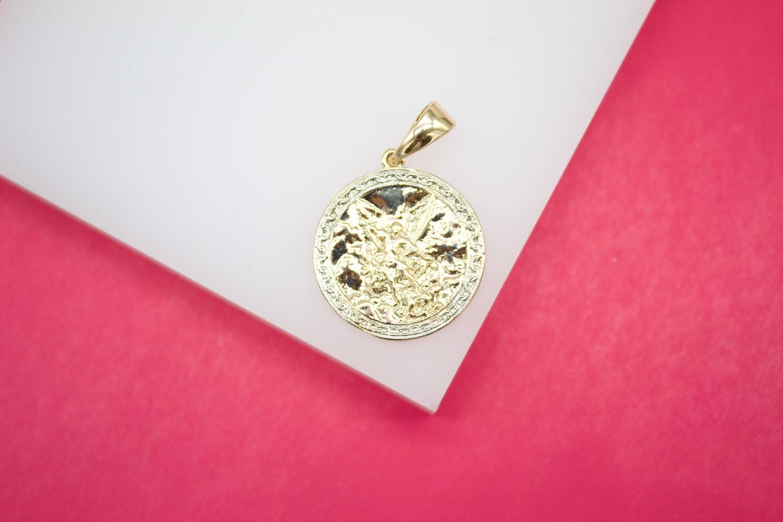 A gold coin pendant featuring a detailed depiction of Saint Michael the Archangel rests on a white display surface against a vibrant pink background.