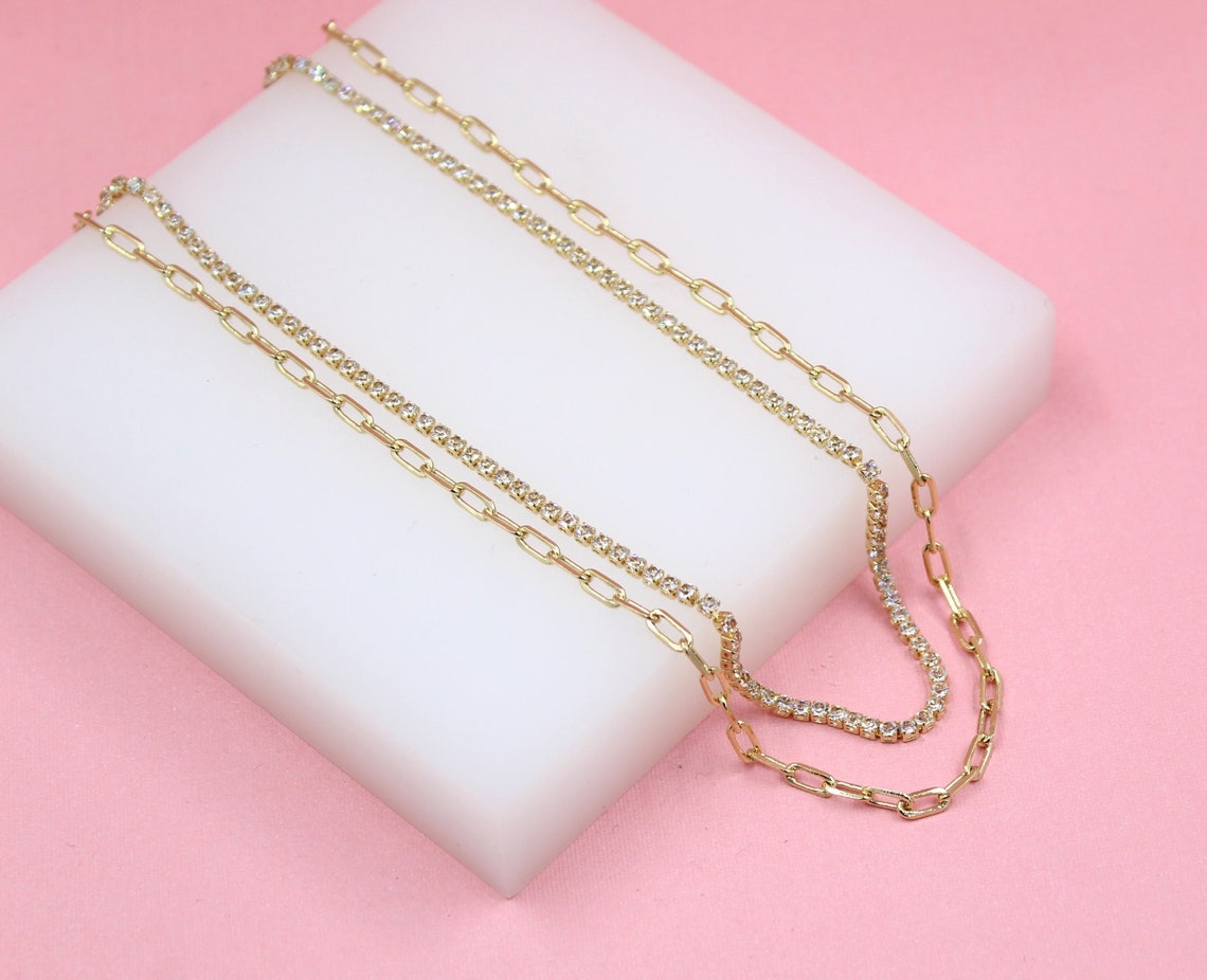 A layered gold chain necklace features a delicate paperclip link strand paired with a sparkling rhinestone tennis chain, displayed on a white block against a soft pink background.