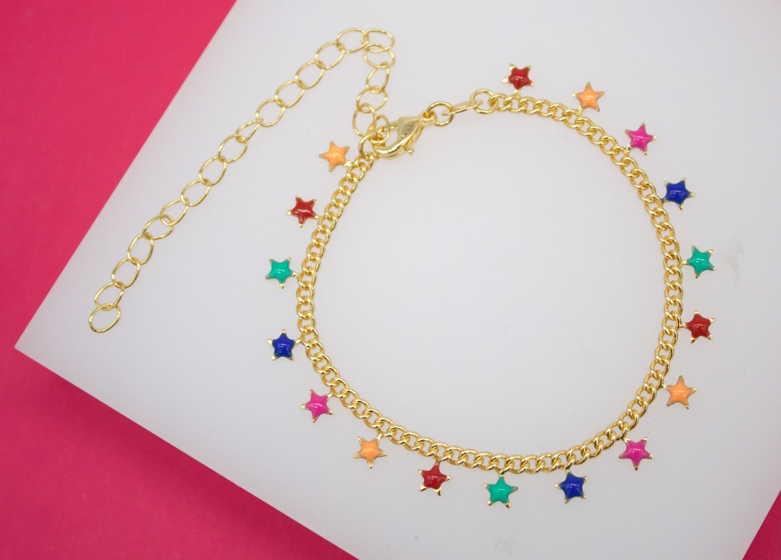 A gold chain charm bracelet dangles colorful enameled star pendants against a white and pink background.