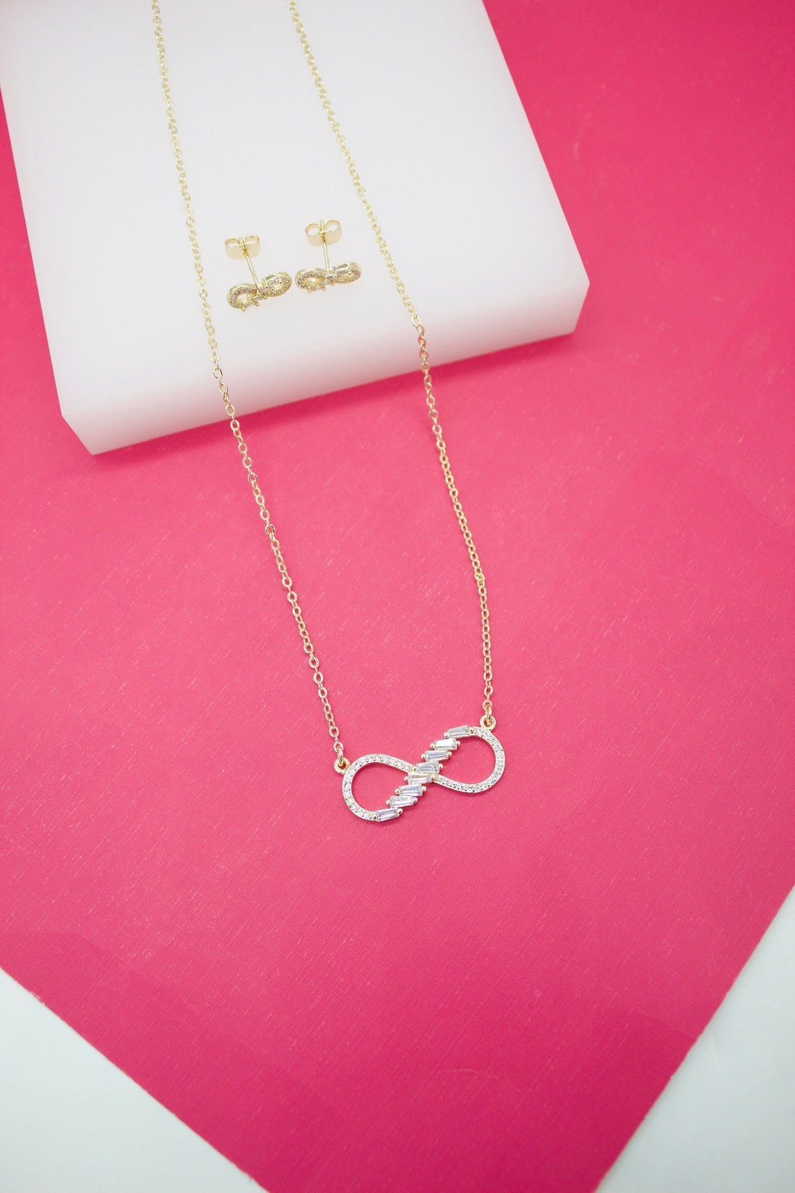 A delicate gold infinity necklace with sparkling cubic zirconia stones rests on a bright pink surface alongside matching ribbon-shaped stud earrings.