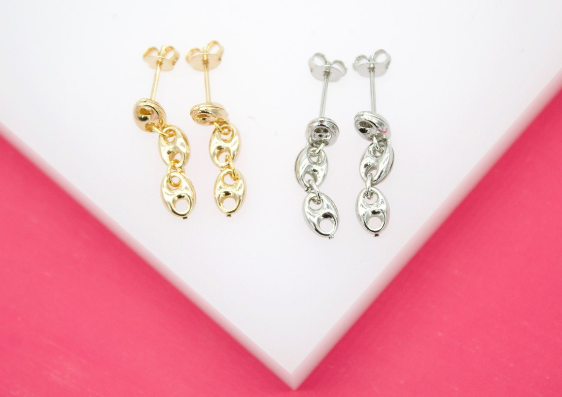A pair of gold and a pair of silver mariner chain drop earrings lie on a white display against a pink background.