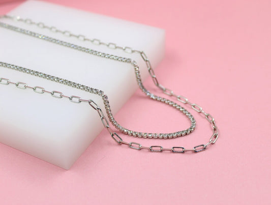 Layered silver necklaces featuring delicate link chains and sparkling diamond tennis chains rest gracefully on a white display block against a soft pink background.
