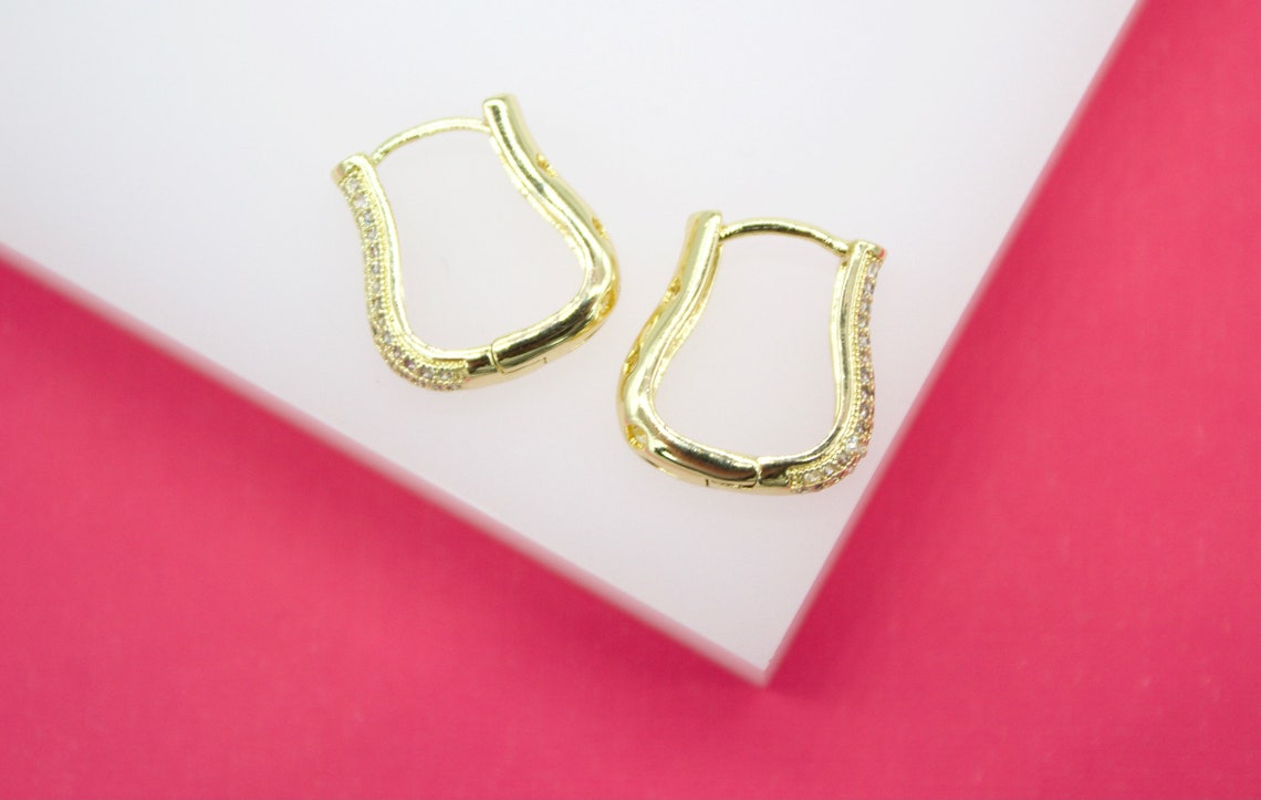 A pair of geometric gold huggie hoop earrings featuring pavé diamond accents rests on a white and pink display surface.
