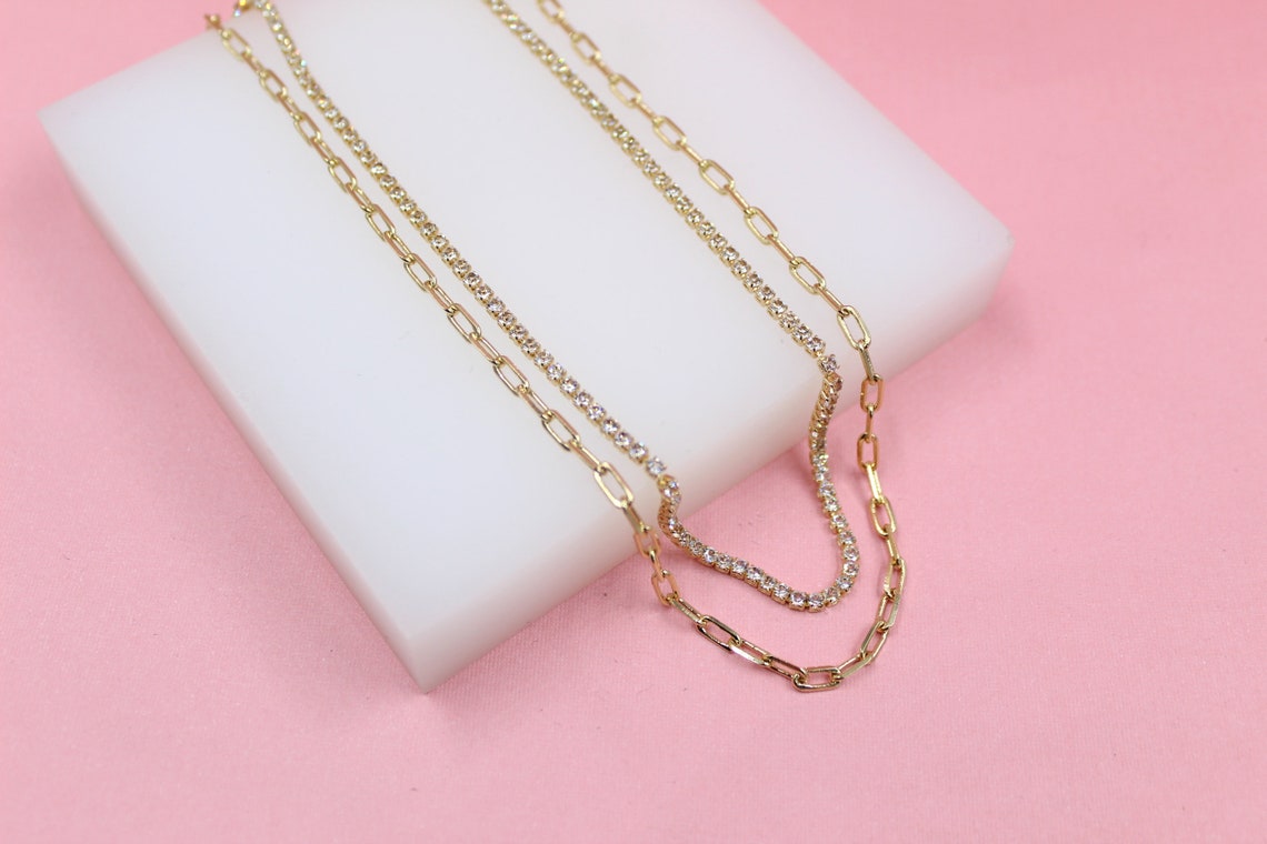 A layered gold necklace set featuring a sparkling diamond tennis chain and a matching link chain rests on a white display block.