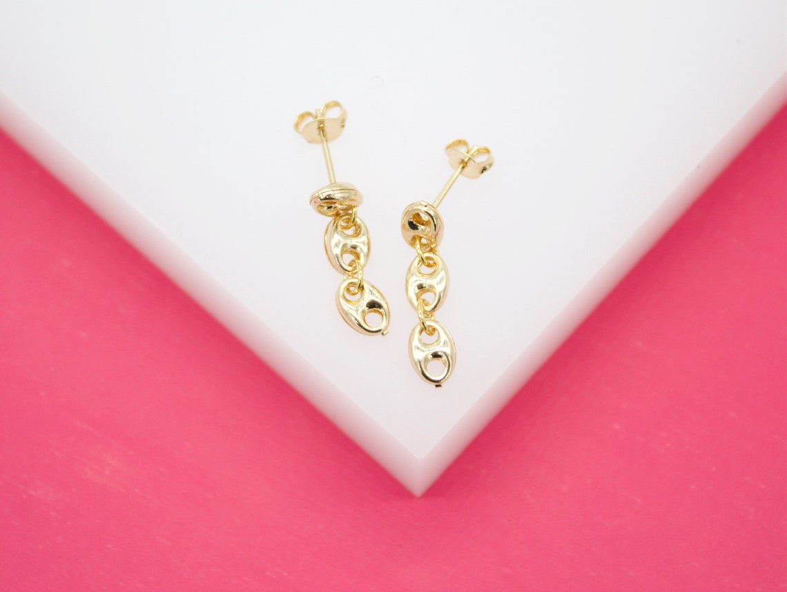 A pair of polished gold mariner link drop earrings rests against a white and pink display stand.