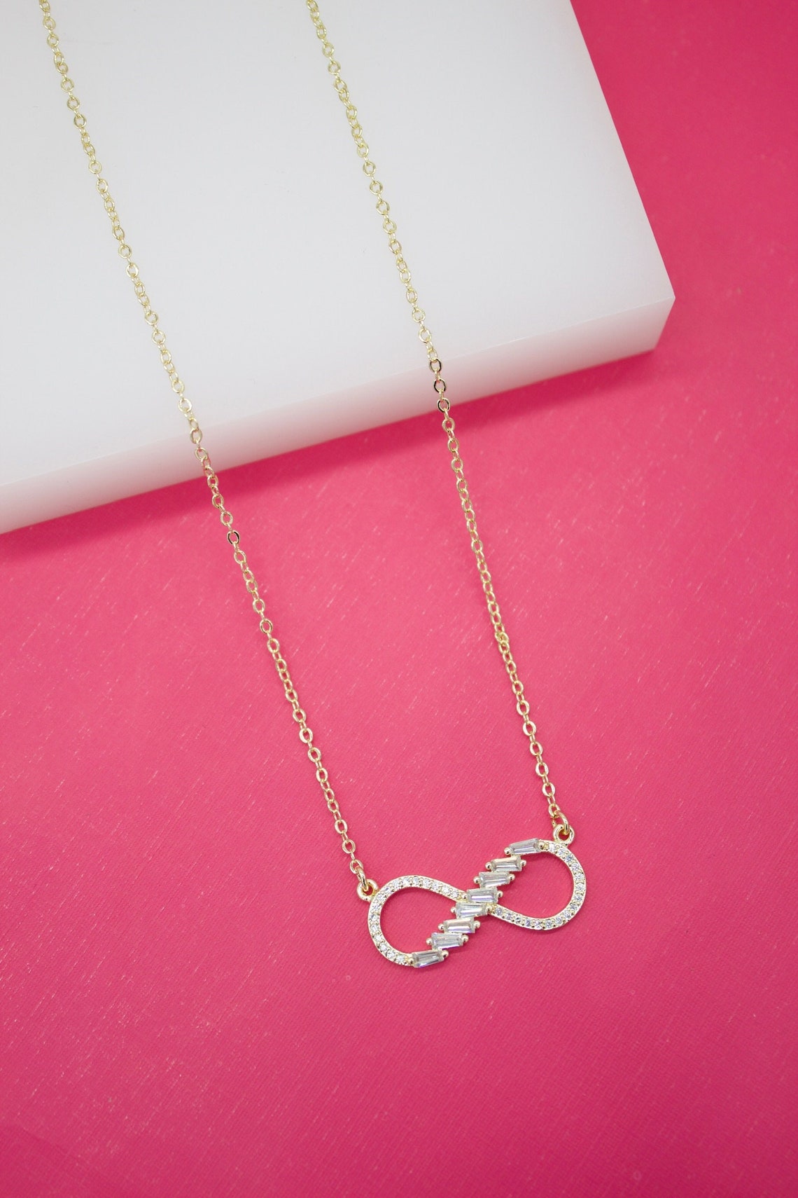 A gold infinity necklace featuring sparkling cubic zirconia stones rests against a vibrant pink backdrop.