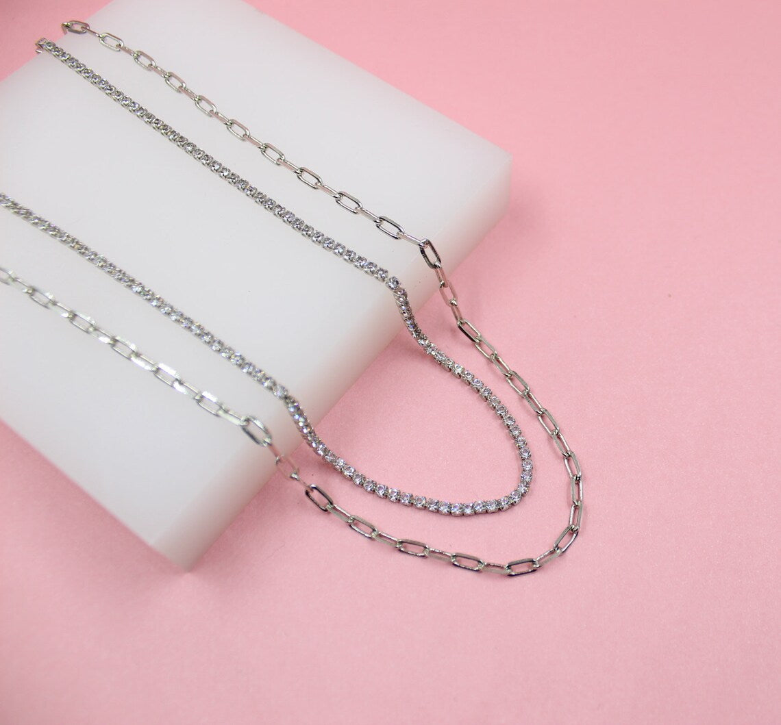 Four layered silver necklaces, including two paperclip chains and a sparkling diamond tennis chain, rest on a white display block against a soft pink background.