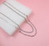 Four layered silver necklaces, including two paperclip chains and a sparkling diamond tennis chain, rest on a white display block against a soft pink background.