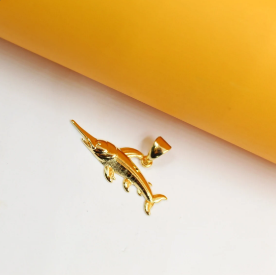 A shiny gold swordfish pendant rests against a white surface beneath a warm yellow backdrop.