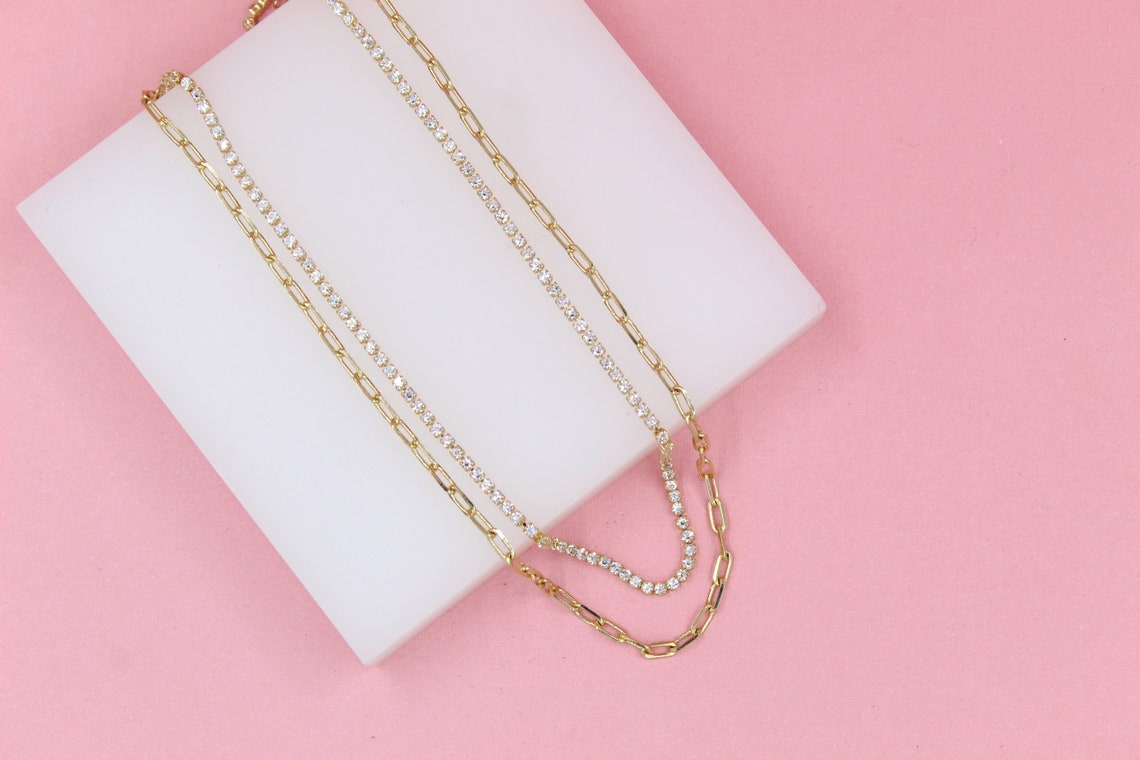 Two layered gold necklaces featuring a dainty link chain and a sparkling rhinestone tennis chain rest gracefully on a white display block against a vibrant pink background.