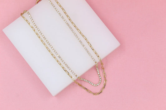 Two delicate gold chain necklaces, one featuring sparkling rhinestones and the other a link design, rest gracefully on a white display block against a vibrant pink background.