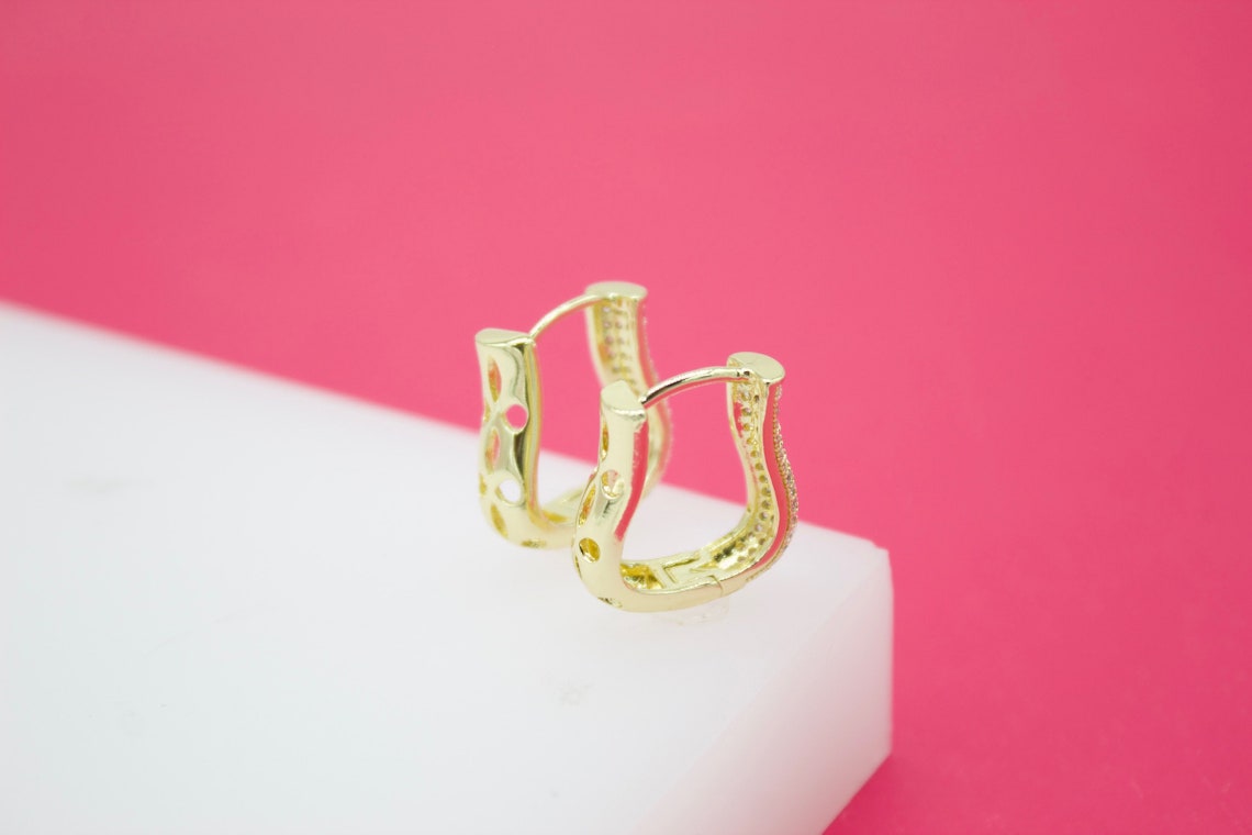 A pair of ornate gold hoop earrings resting on a white display block against a pink background.