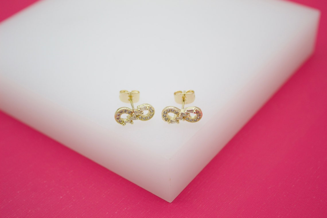 A pair of gold infinity symbol stud earrings encrusted with sparkling zirconia stones rests on a white display block against a vibrant pink background.