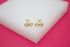 A pair of gold infinity symbol stud earrings encrusted with sparkling zirconia stones rests on a white display block against a vibrant pink background.