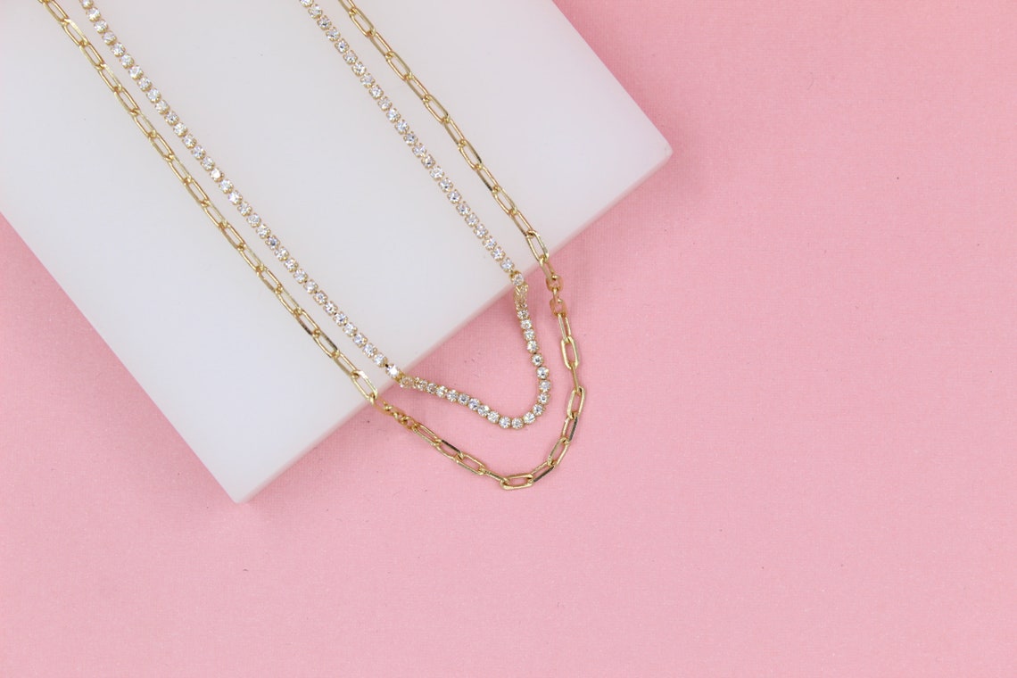 A sparkling diamond tennis necklace and a gold paperclip chain necklace rest draped over a white display block against a soft pink background.