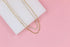Two layered gold necklaces featuring a dainty paperclip chain and a sparkling rhinestone tennis chain rest on a white display against a pink background.