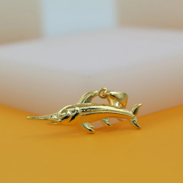 A polished gold swordfish pendant rests on an orange surface against a neutral background.