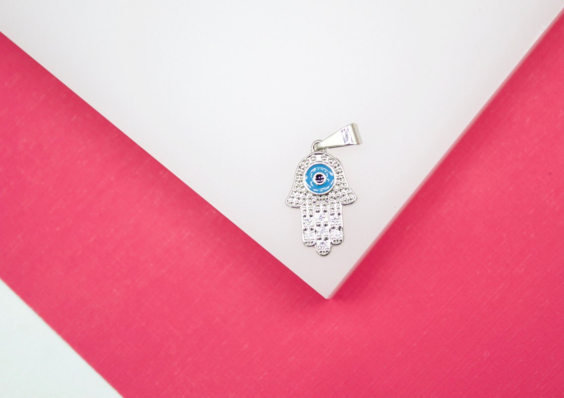 A sparkling silver hamsa hand pendant with a blue evil eye centerpiece rests on a white and pink background.