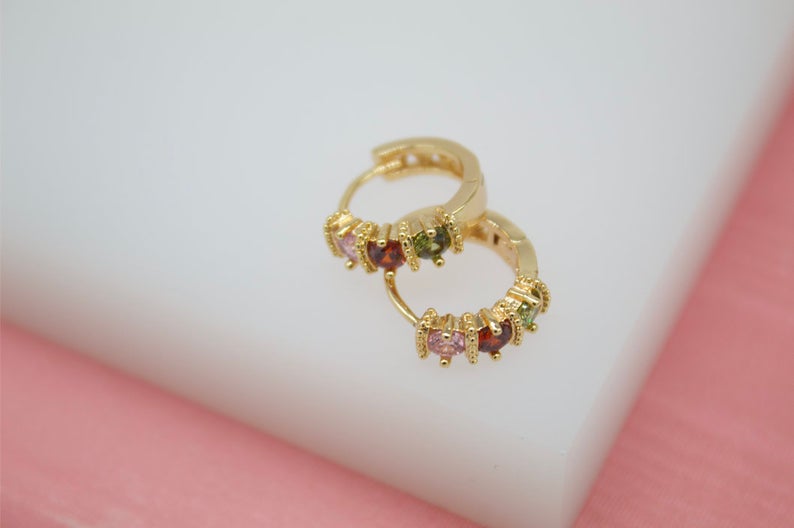 A pair of gold huggie hoop earrings with multicolored crystal stones rests on a white display surface.