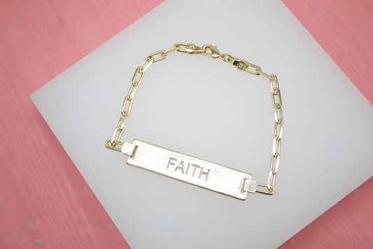 A gold ID bracelet with the word FAITH engraved on the rectangular bar rests on a white display pad against a pink background.