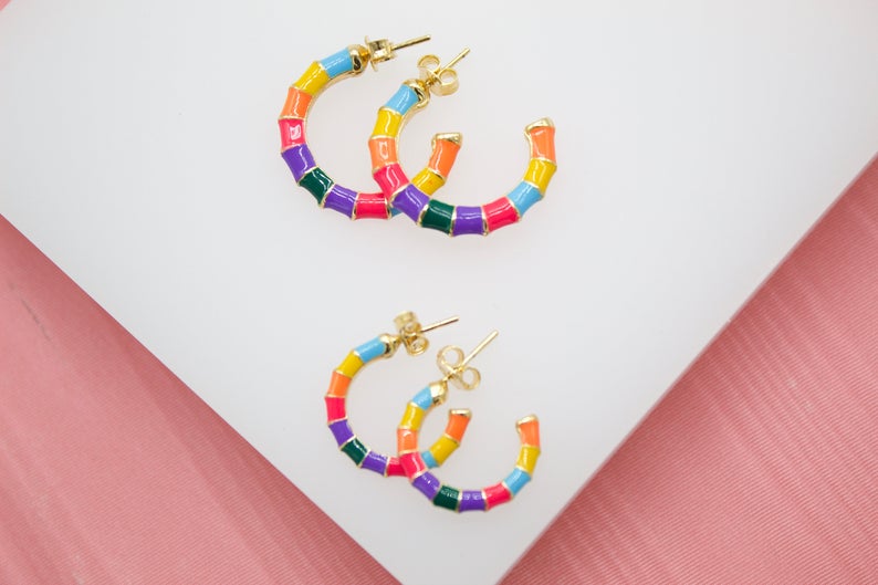 Two pairs of colorful enamel bamboo hoop earrings rest on a white surface against a pink background.