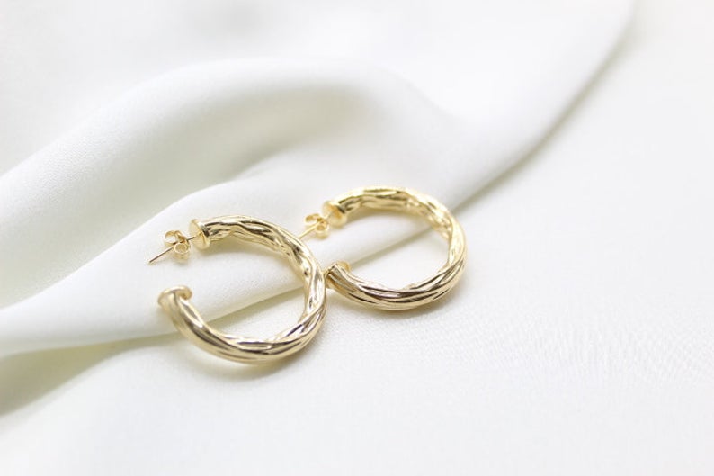 A pair of textured gold hoop earrings rests elegantly on a soft white silk fabric.